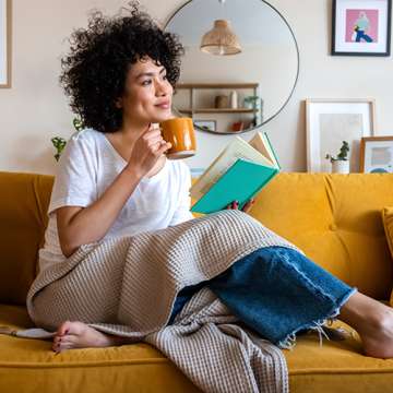 female presenting person sipping a drink on a couch while reading a book