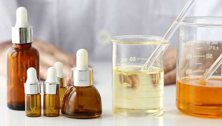 five small dark brown bottles used for essential oils on the left with two large beakers with essential oils inside, one a white table with a white man in a lab coat in the background