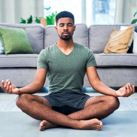 dark skinned, male presenting person sitting in living room meditating