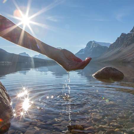 light-skinned hand and forearm scooping water from a clear lake with mountains in the background
