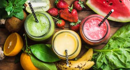 three different color smoothies in glasses with straws, green, yellow, red. surrounded by fruits and vegetables such as spinach, bananas, oranges, strawberries and more. 