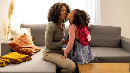 two female presenting people of dark skin tone, one presumably the mom and the other a young daughter with a backpack on hugging on a couch 