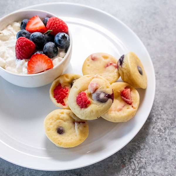 six protein pancake bites on a white plate along with a cup of mixed berries and yogurt