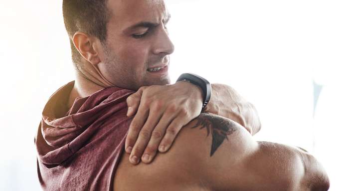 muscular presenting as male in sleeveless workout shirt reaching with left hand to massage right shoulder grimacing