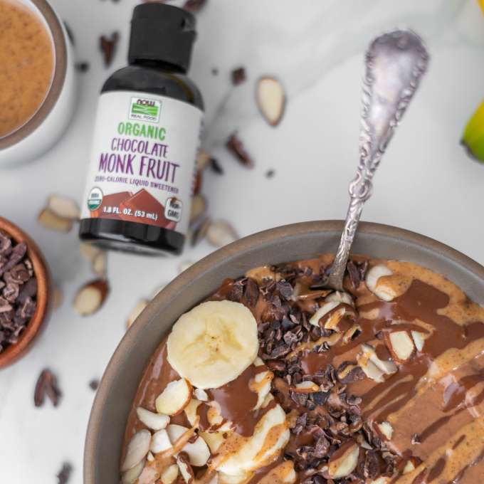 brown bowl filled with double chocolate smoothie bowl mixture including sliced bananas on top and a bottle of NOW Chocolate Monk Fruit sweetener laying to the top left