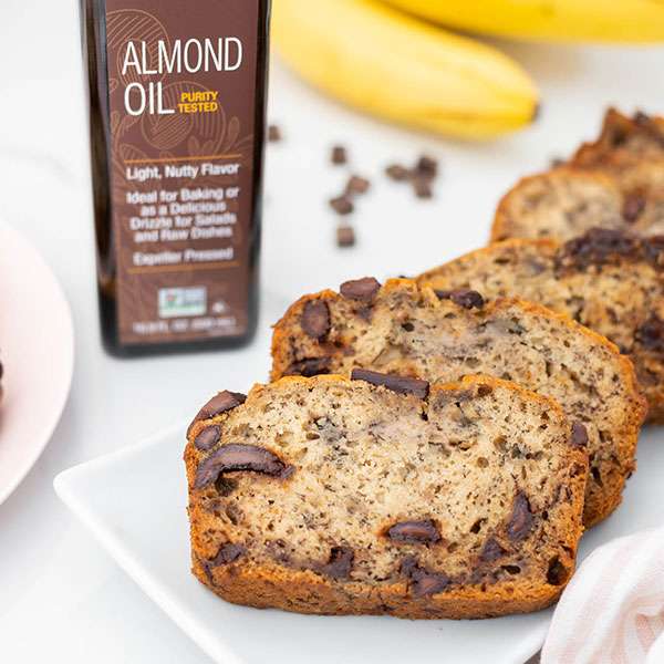 chocolate banana bread slices layed out on a white serving plate, a bottle of NOW Ellyndale Almond Oil to the left of the slices and bananas and chocolate chips in the background