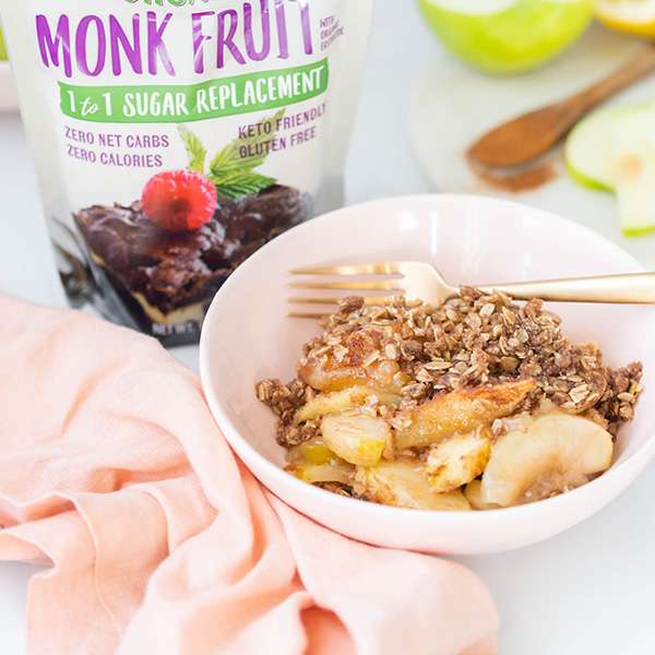 spiced apple crisp in a light pink bowl with a small scoop of ice cream on top and a bag of NOW Monk Fruit Sweetener to the left of the bowl