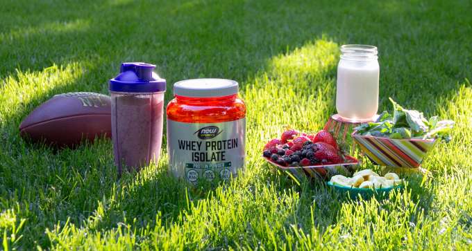 grouping of items sitting in the grass. Football, smoothie in a NOW Shaker Bottle, NOW Sports Whey Protein Isolate, Bowl of berries, bowl of spinach, bowl of banana's, glass jar of milk