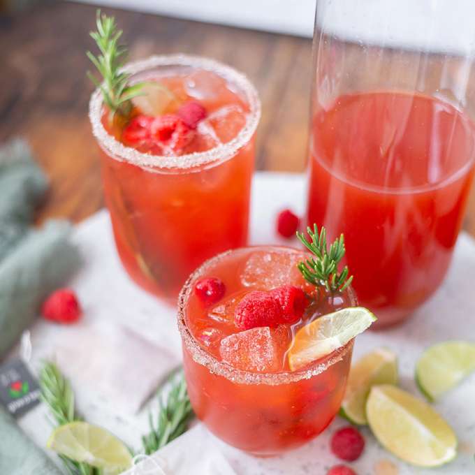 two clear glasses filled with red iced tea with sugar around the rim and a green sprig rising from the top, and a pitcher half filled with same red tea
