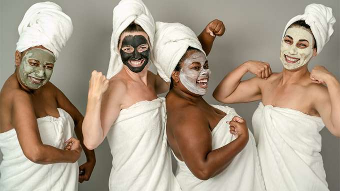 four female presenting people of various races in white towels flexing their biceps with face masks on