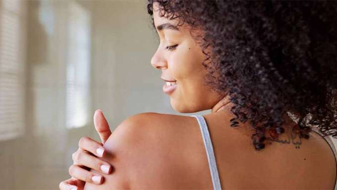 medium toned skin female presenting person showing her back and rubbing in a lotion on her shoulder