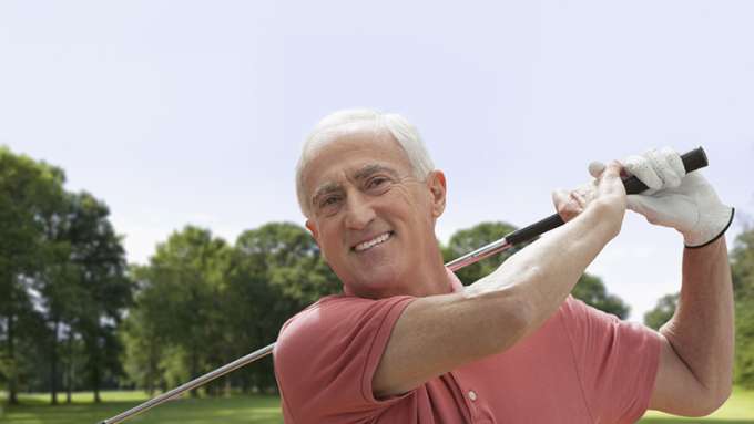 A male-presenting, light-skin person with grey hair smiles during a swing at an outdoor golf course.