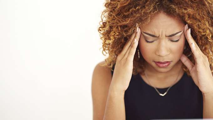 A female-presenting person with dark skin and brown curly hair rubbing her temples and grimacing.