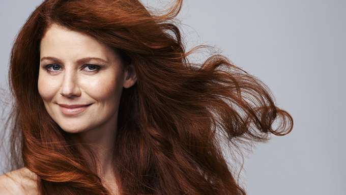 A female-presenting person with light skin, long red hair and blue eyes looks at the camera