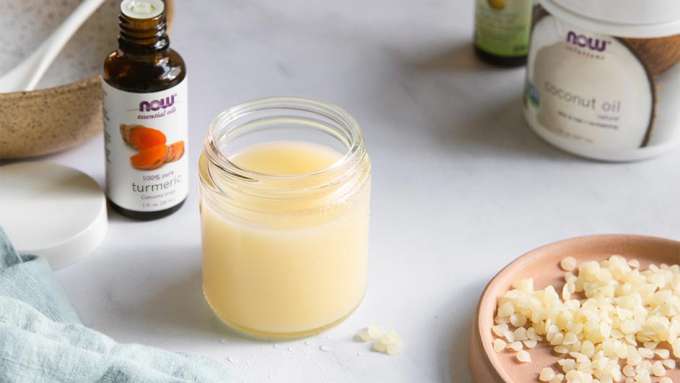 glass jar filled with Turmeric Oil Cleanser, Bottle of NOW Turmeric Essential Oil, NOW Solutions Coconut Oil, and beeswax pellets
