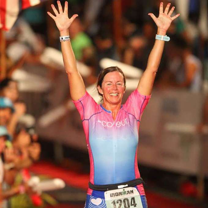 Heather Casey has her brown hair pulled back and is wearing a t-shirt with red on the upper one third and blue on the lower one third and a race number at her waist. Her hands are in the air with all ten fingers fanned out and she is smiling after a race.