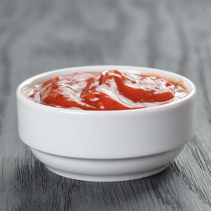 A small dipping bowl on a black wooden table is filled with bright-red Sugar-Free Ketchup. 
