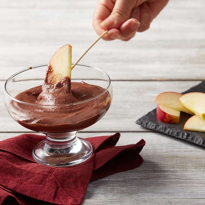 An apple slice on a stick is dipped into a short glass filled with Coconut Cacao Dipping Sauce