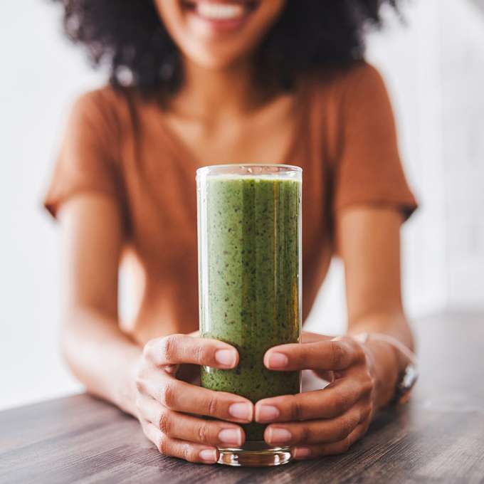 dark skinned female presenting person holding a glass of green smoothie. 