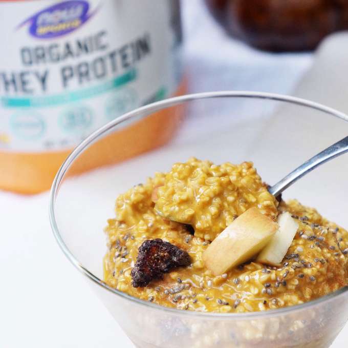 Round glass bowl of cooked steel cut oats with a spoon in the bowl and the handle of it sticking out on the right, topped with chia seeds, dried fig bits and apple chunks. On the white counter in the blurred background from left to right is a NOW Sports bottle of whey protein and the bottom portion of a brown ceramic pumpkin.