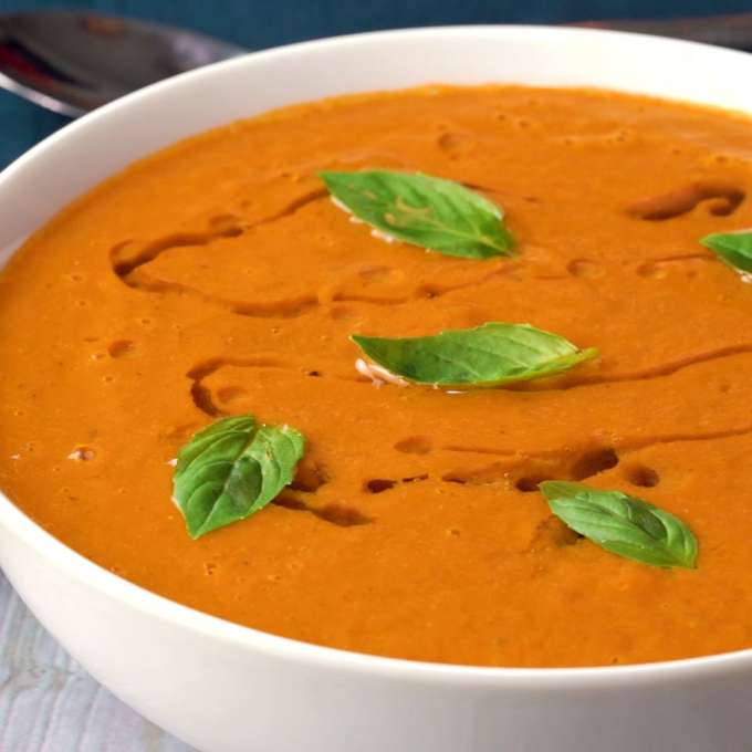 White ceramic round bowl of tomato soup dark pumpkin orange in color with a drizzle of coconut oil and five small basil leaves on top.
