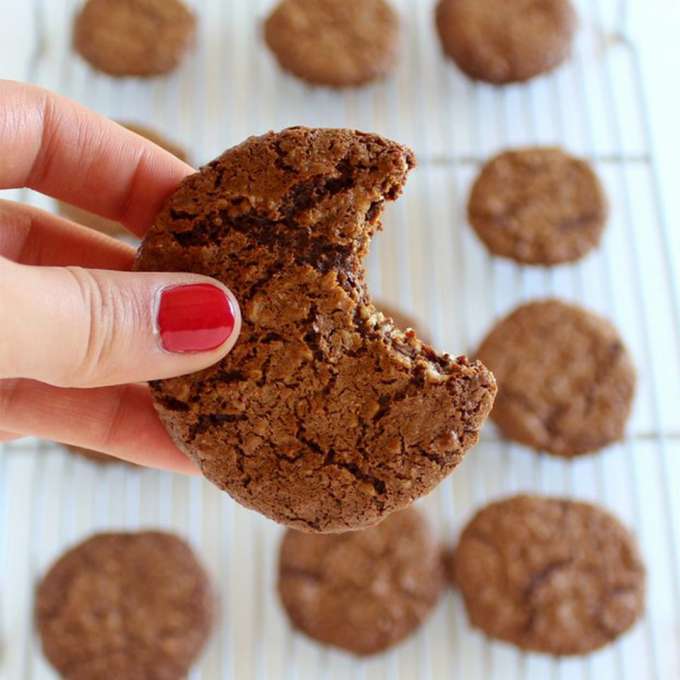 Light skin hand with red nail polish holding a cookie with a bite out of it. Background is a cooling tray with cookies on them on a white countertop. The cookies are medium brown in color.