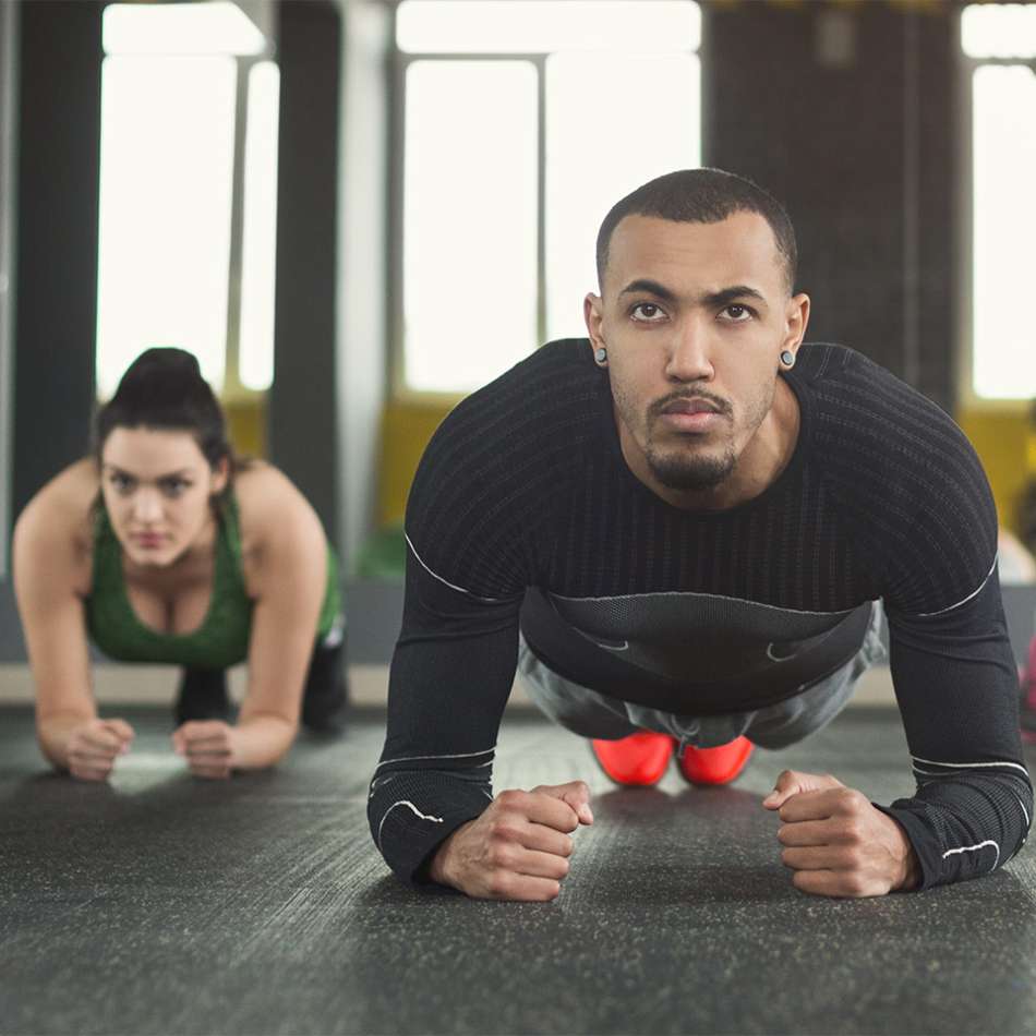male presenting on the right in workout clothes laying on a mat propped up on his forearms facing the camera and a female presenting person on the left in the same position