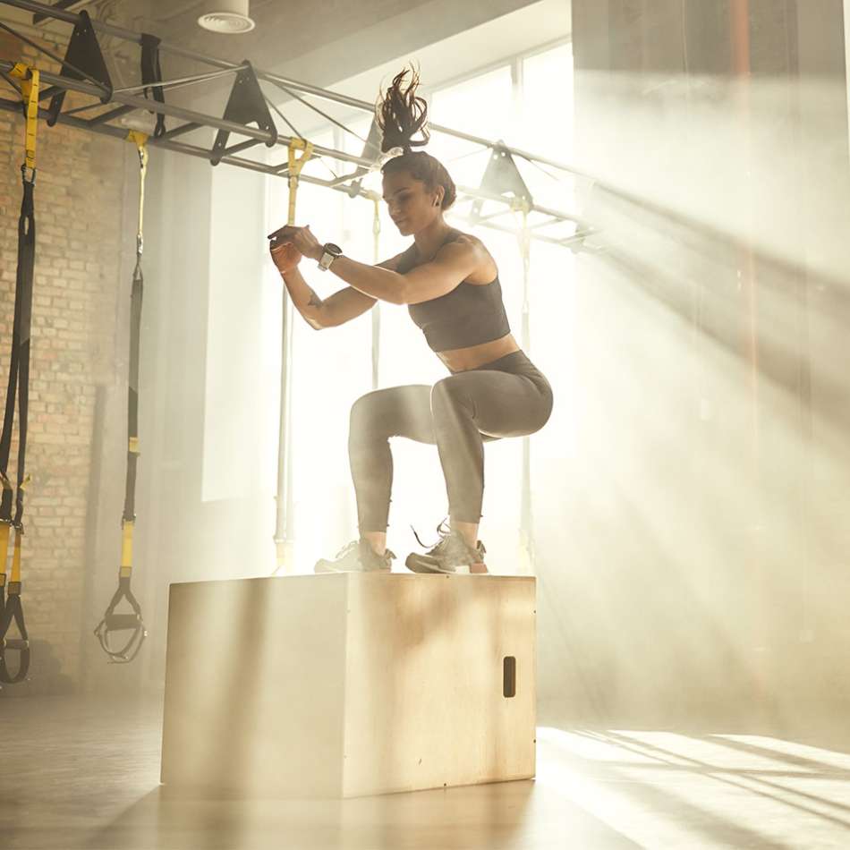 presenting as woman in workout clothes jumping onto a platform in a sunlit gym