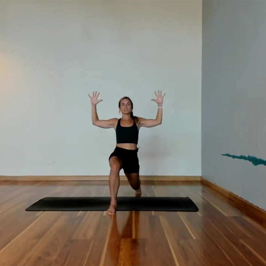 female presenting in black workout tights with hands raised and lunging forward; wood floor with yoga mat
