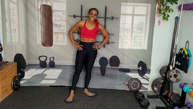 De Bolton standing with her hands on her hips in her home gym wearing a bright red top and black pants