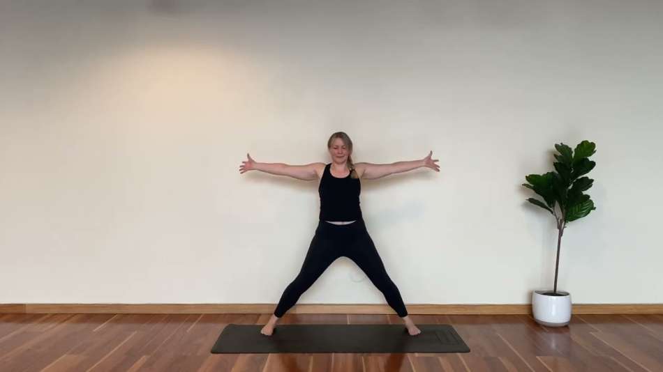 light skin female presenting person in all black work out clothes standing on black yoga mat with her arms stretched wide