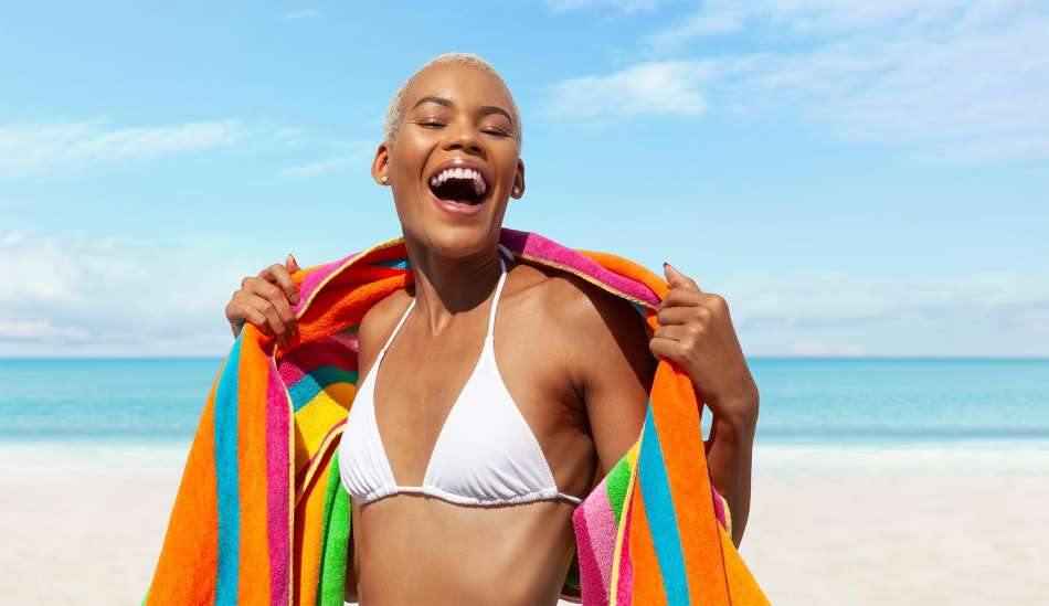 dark skinned woman in a white bikini top smiling holding onto a colorful towel around her shoulders